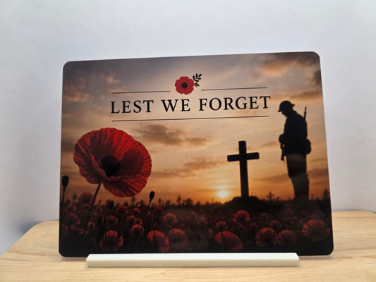 Lest We Forget remembrance metal sign with red poppies, a soldier silhouette and memorial cross at sunset, displayed on a white stand.