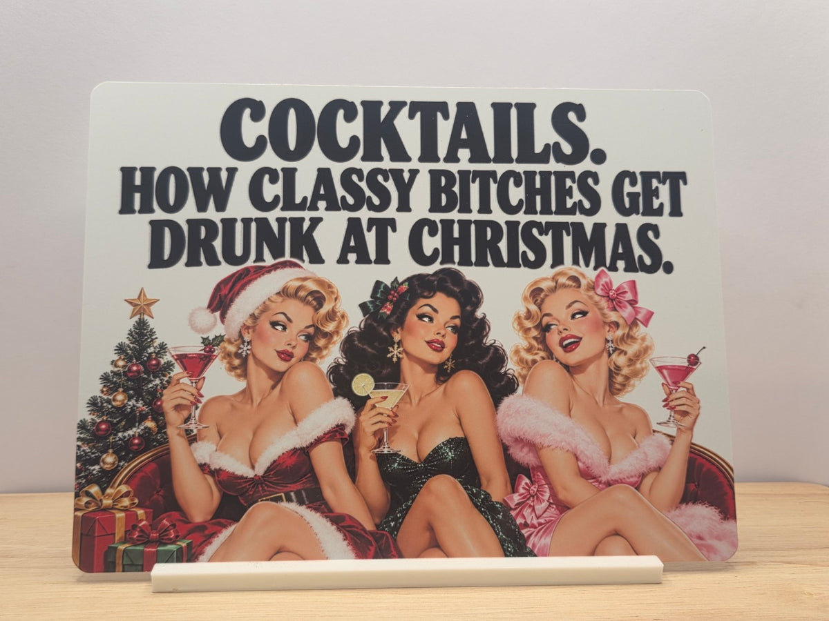 Cocktails Christmas humour metal sign with three retro women holding drinks and bold black lettering, displayed in a white stand