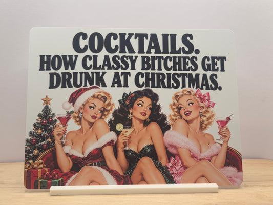 Cocktails Christmas humour metal sign with three retro women holding drinks and bold black lettering, displayed in a white stand
