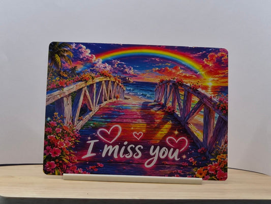 I miss you metal sign with a rainbow over an ocean sunset and a flower-lined bridge, displayed in a white stand
