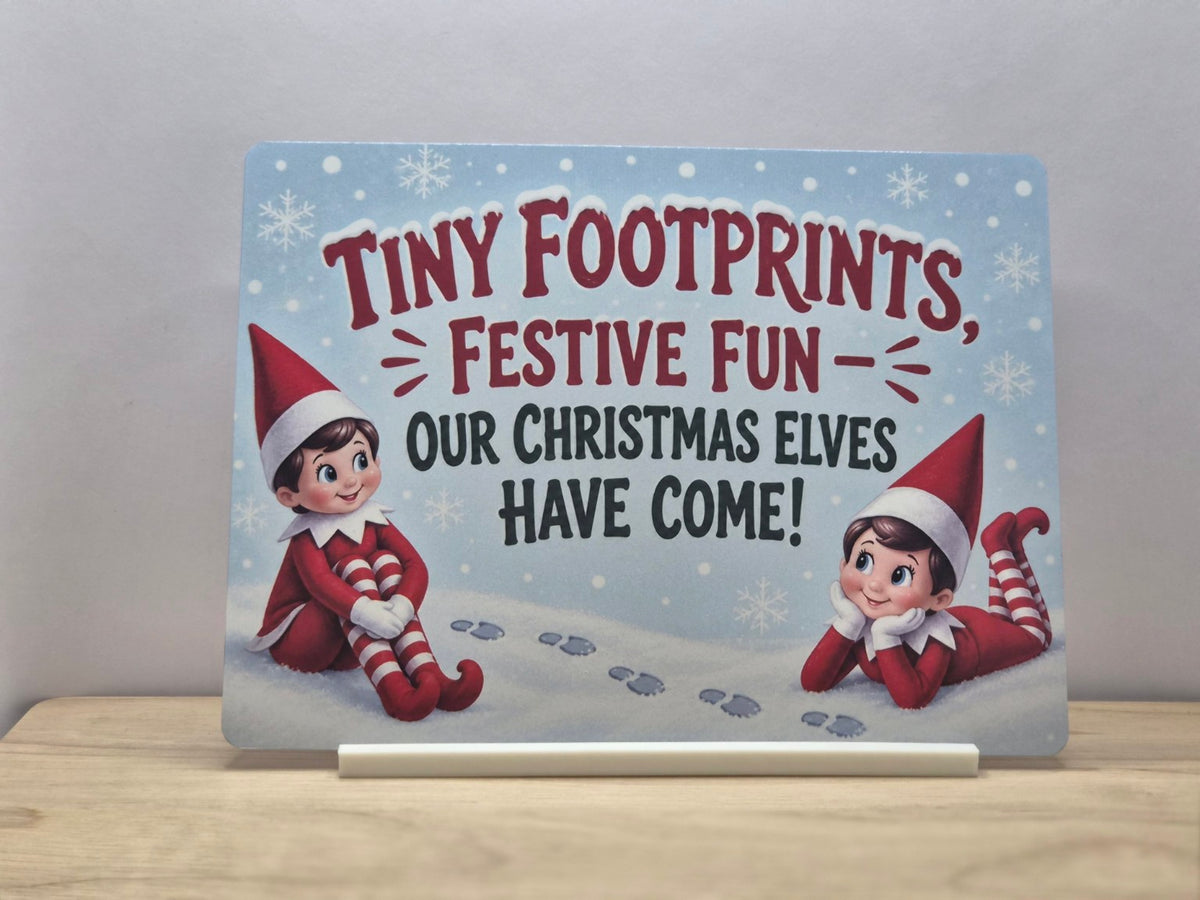 Our Christmas Elves Have Come metal sign with two red-clad elves, footprints and snowflakes, displayed in a white stand