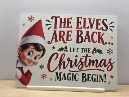 The Elves Are Back metal sign with a smiling elf, red festive lettering, holly and snowflakes, displayed in a white stand