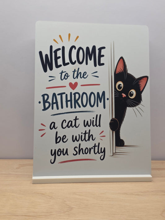 Welcome to the Bathroom metal sign with a black cat peeking around the lettering, displayed in a white stand